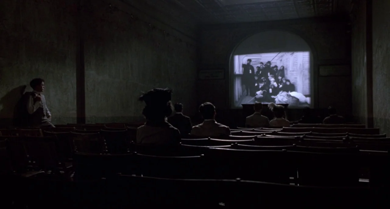 Chaplin (1992) movie still: wide — A dark, sparsely occupied early 20th-century theater features rows of empty wooden seating. Several people sit…