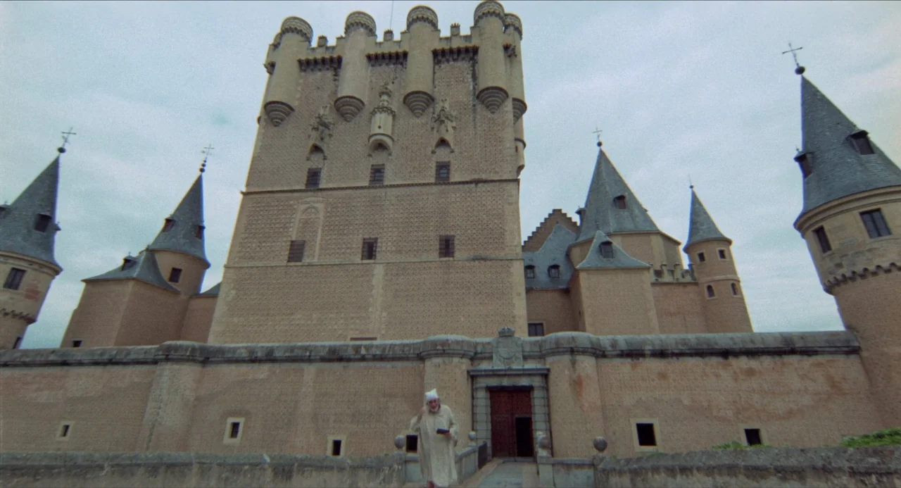 The Three Musketeers (1973) movie still: wide — A massive medieval stone castle with tall towers rises against an overcast, cloudy sky. A solitary…