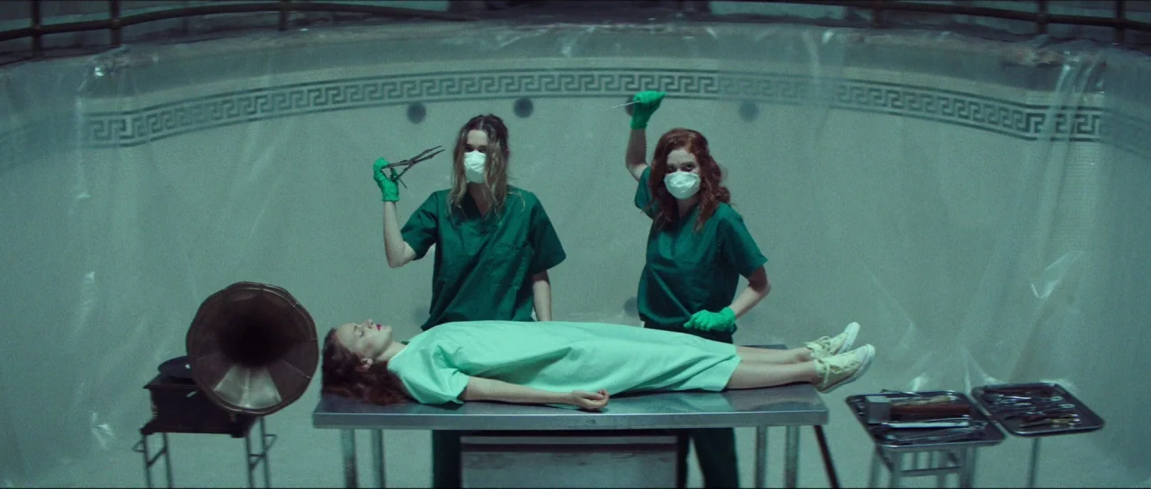 Braid (2018) movie still: wide — A drained tiled pool with plastic sheeting against the curved walls. Two figures in scrubs stand behind a patient…