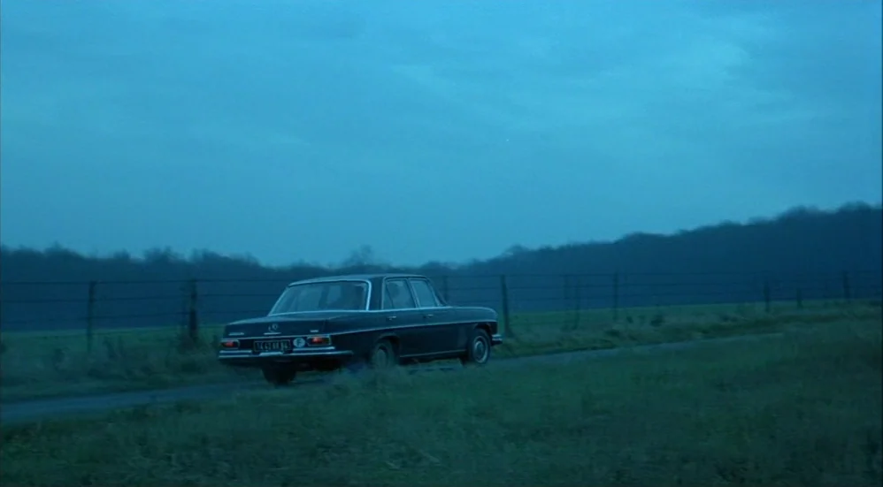 Un Flic (1972) movie still: wide — A dark vintage sedan travels along a narrow asphalt road bordered by a wire fence. The scene is set in an open…