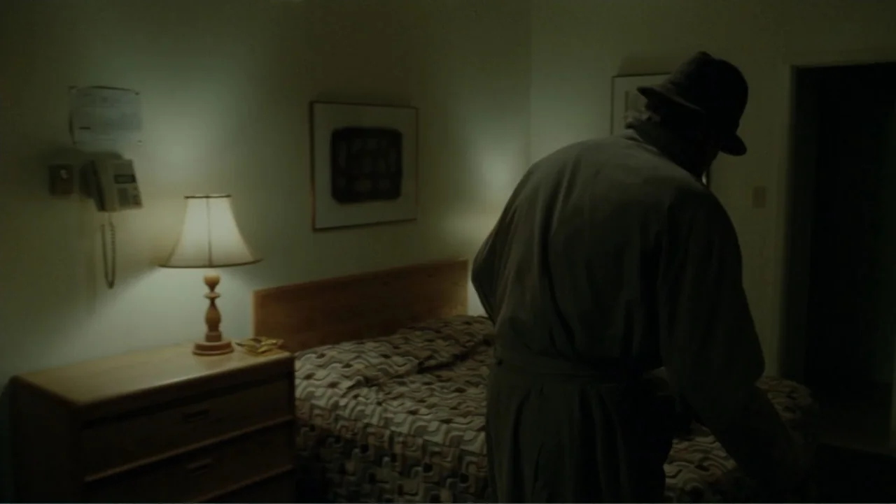 The Words (2012) movie still: medium — A dimly lit motel room contains a bed, a dresser, and a lamp. A man wearing a trench coat and hat stands near…