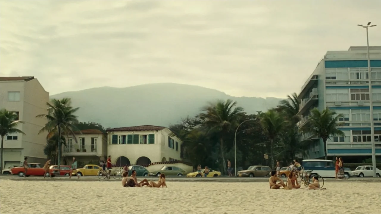 I'm Still Here (2024) movie still: wide — A wide view of a sandy shoreline with palm trees and mid-century architecture in background. Small groups…