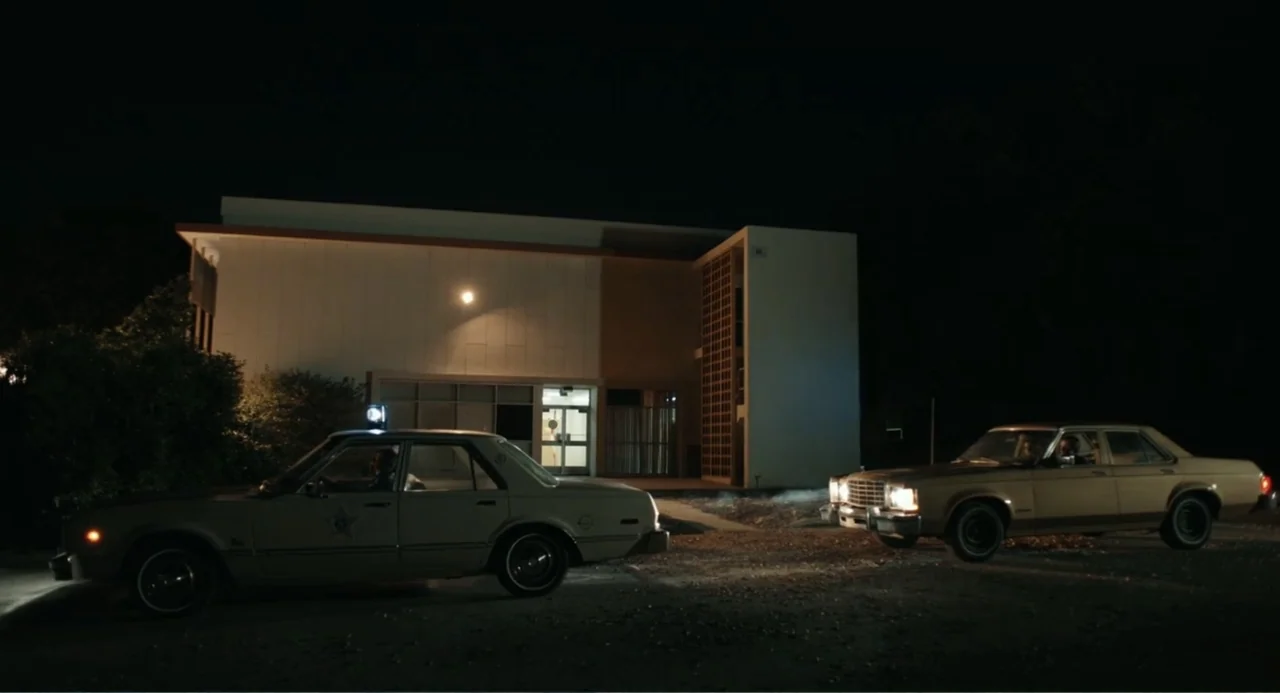 Ted Bundy: American Boogeyman (2021) movie still: wide — A low-slung, modern building sits at night behind two parked sedan vehicles. The area is…