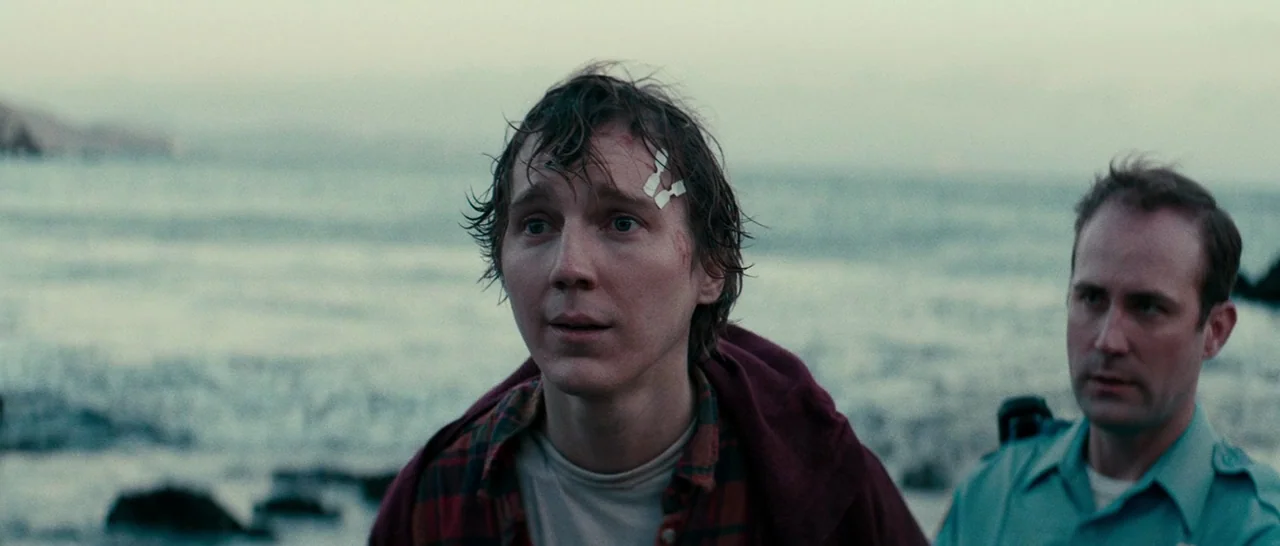 Swiss Army Man (2016) movie still: medium — Two men stand on a rocky coastline during the twilight hour. The younger man appears disheveled while an…