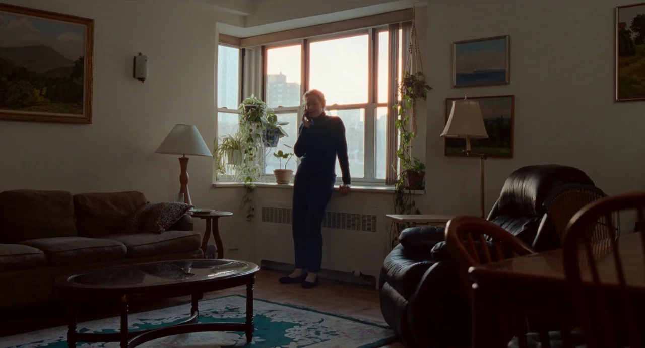 His Three Daughters (2023) movie still: wide — A living room interior featuring a sofa, coffee table, and a large window. A woman stands by the…