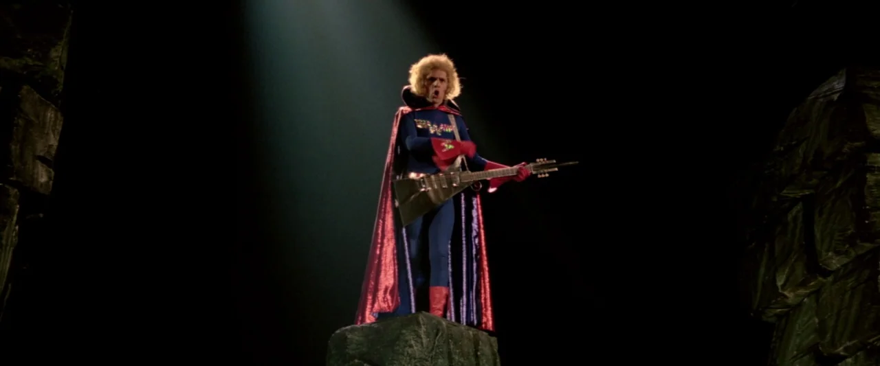 Lisztomania (1975) movie still: fullbody — A man in a superhero costume stands atop a jagged rock in darkness. He holds a V-shaped electric guitar…