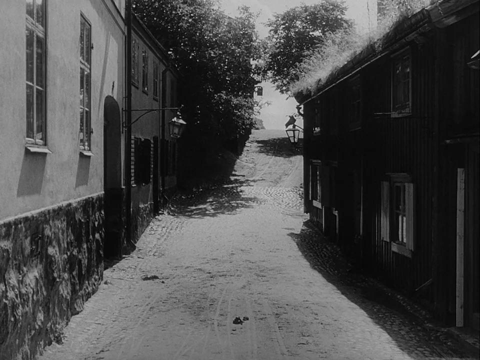The Magician (1958) movie still: establishing — A narrow cobblestone lane passes between old European-style houses with grass-covered rooftops. Lush…