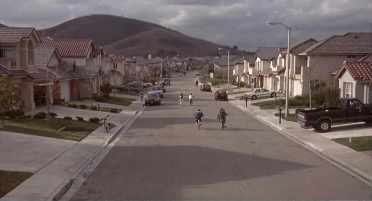 3 Ninjas (1992) movie still: wide — A suburban residential street lined with two-story houses leads toward a distant mountain. Four children ride…