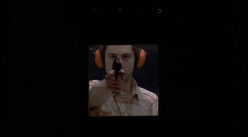 Taxi Driver (1976) movie still: closeup — A man stands in a dark booth at an indoor target practice facility. He stares ahead while aiming a small…