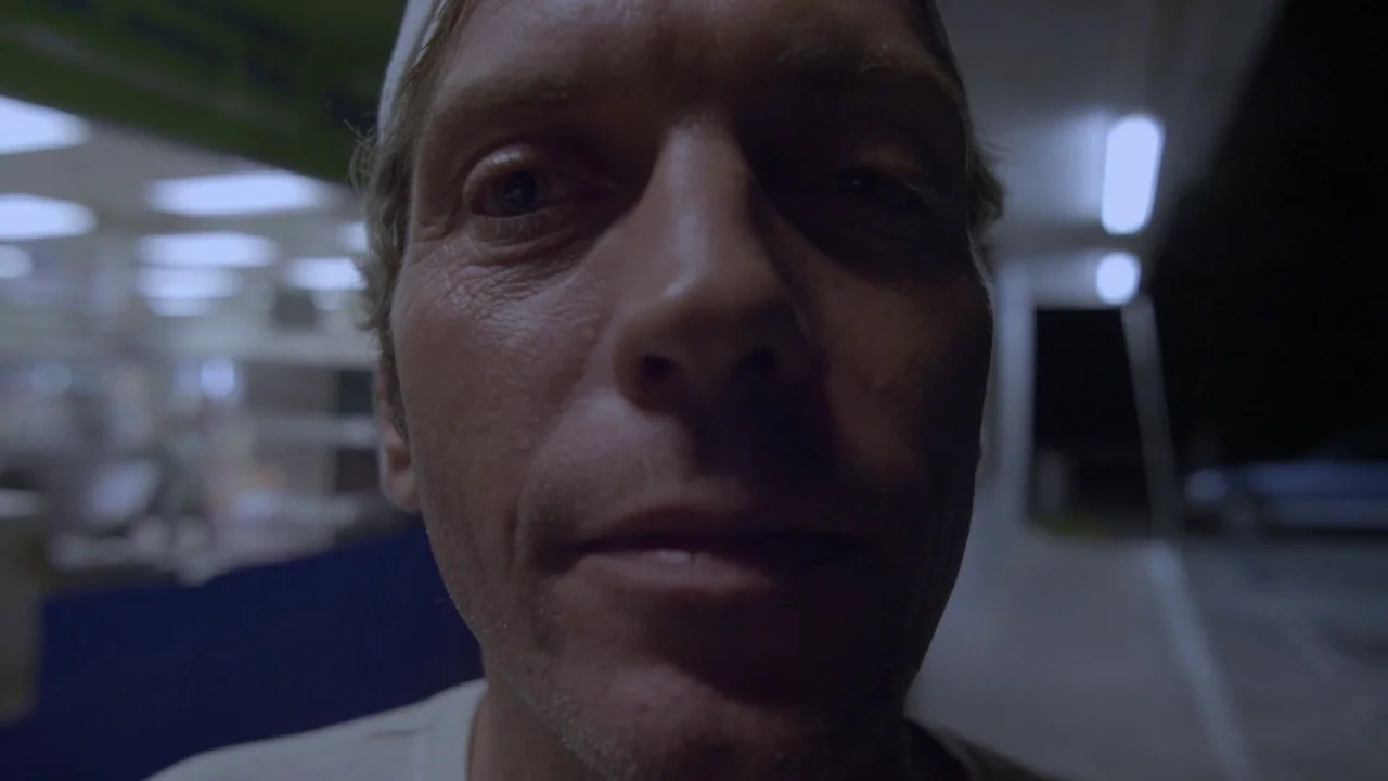 Florida Man (2015) movie still: closeup — A dark outdoor space at night under the fluorescent glow of a gas station. A man stands extremely close to…