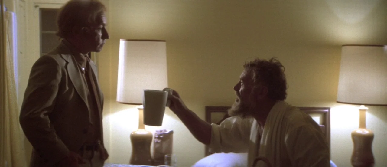 The Long Goodbye (1973) movie still: medium — A dimly lit hotel room with two lamps glowing brightly against a yellow wall. A bearded man in a…