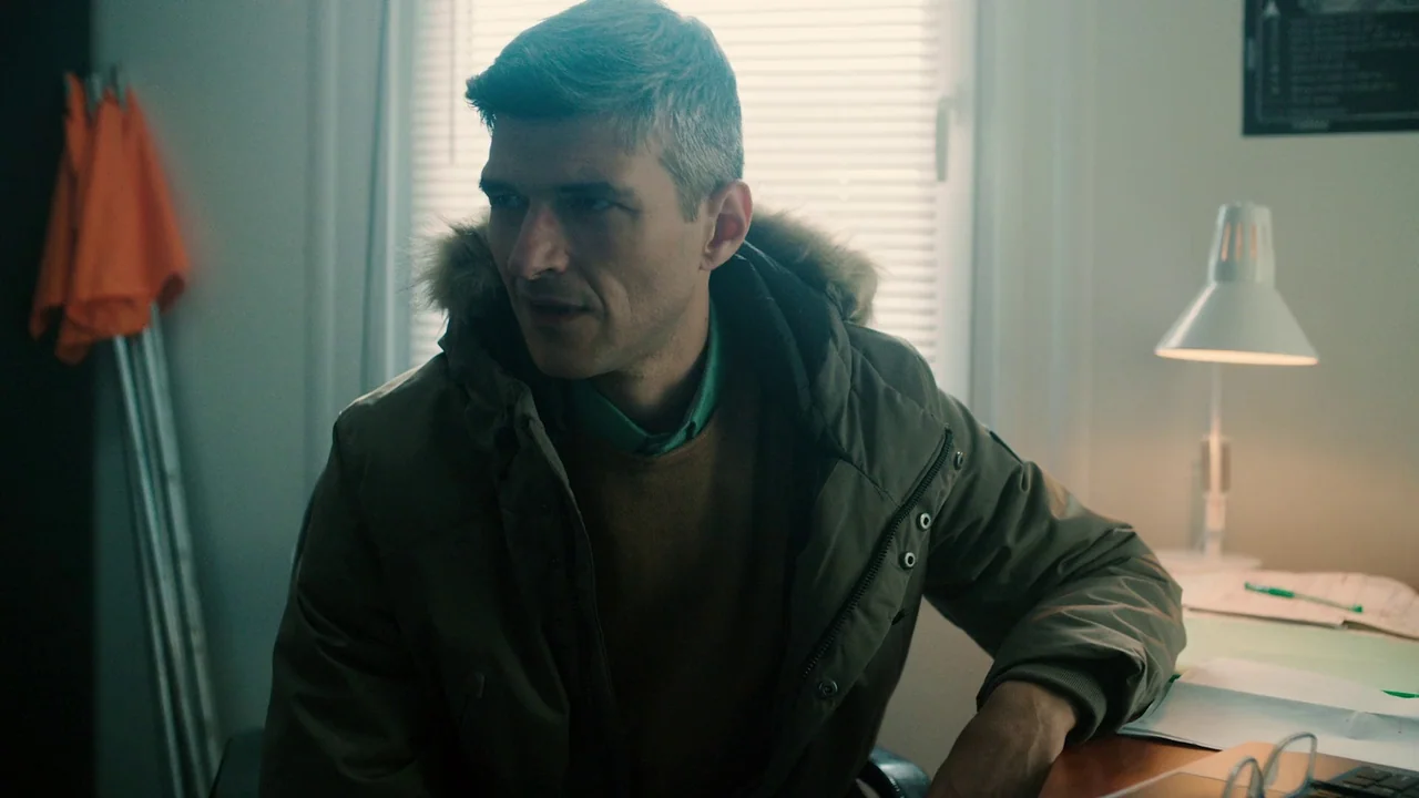 Helll Hole (2024) movie still: medium — A man sits at a desk in a plain room with white Venetian blinds. The workspace includes a lit desk lamp,…