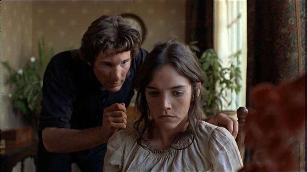 Days Of Heaven (1978) movie still: medium — A furnished interior space with light-colored wallpaper and a window with dark curtains. A man stands…