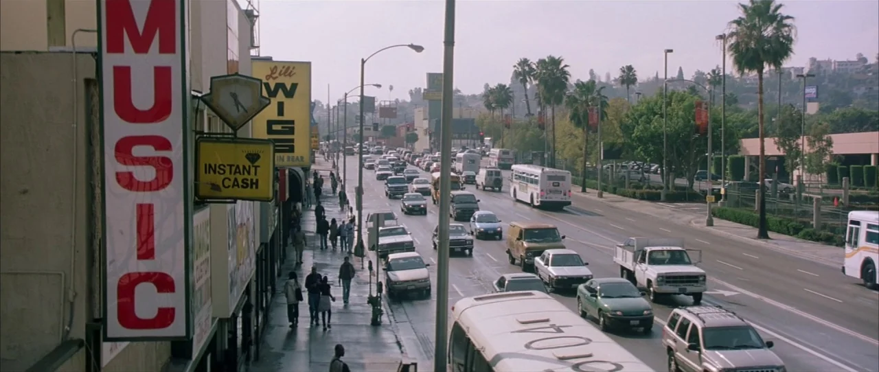 Training Day (2001) movie still: wide — A busy Los Angeles street is filled with various cars and buses during daytime. Pedestrians walk on the…
