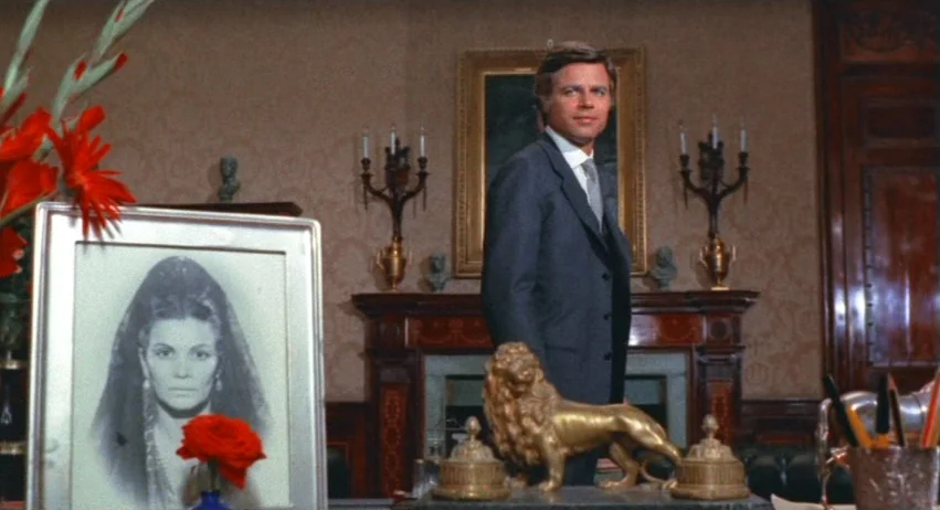 A Lizard In A Woman’s Skin (1971) movie still: medium — A traditional study features patterned wallpaper, a wooden fireplace mantel, and ornate…