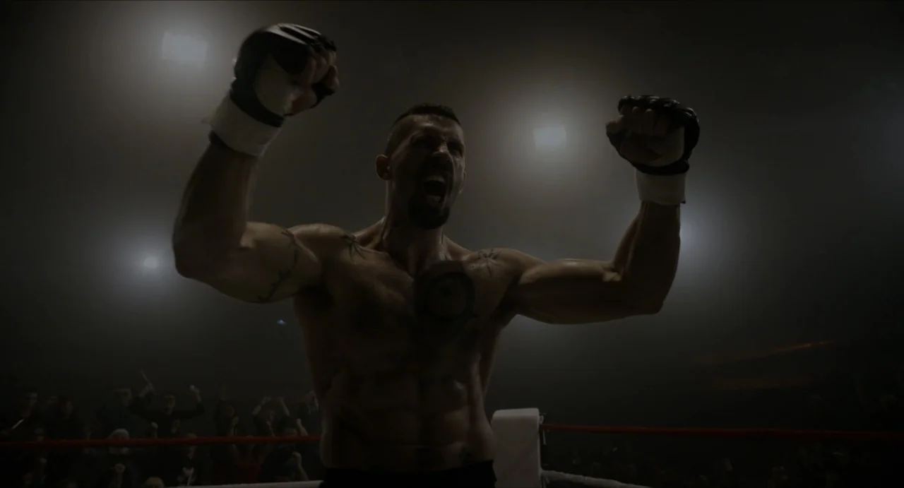 Boyka: Undisputed IV (2016) movie still: medium — A dimly lit combat arena with overhead spotlights shining through a haze of smoke. A bare-chested…