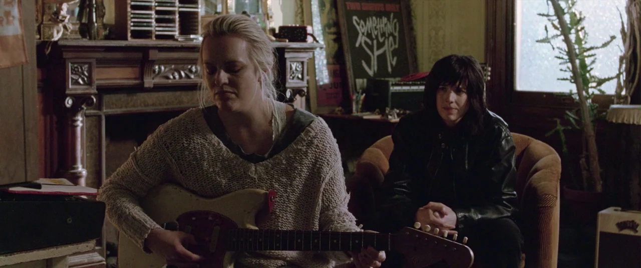 Her Smell (2018) movie still: medium — A cluttered interior space features a brick fireplace, music posters, and houseplants. One woman plays a…