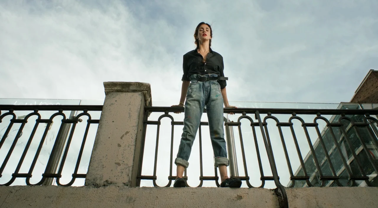 I’m So Excited! (2013) movie still: fullbody — A woman stands on a raised concrete platform behind an ornate metal railing. The sky is partially…