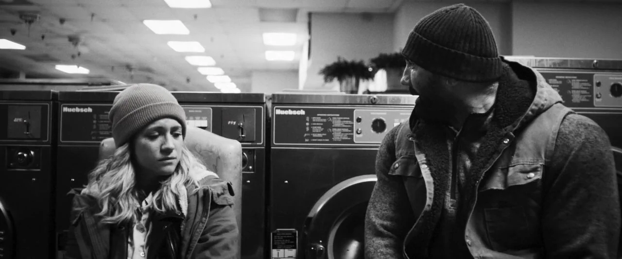 Bushwick (2017) movie still: medium — A row of industrial coin-operated machines sits in a brightly lit, sterile commercial space. Two people sit on…