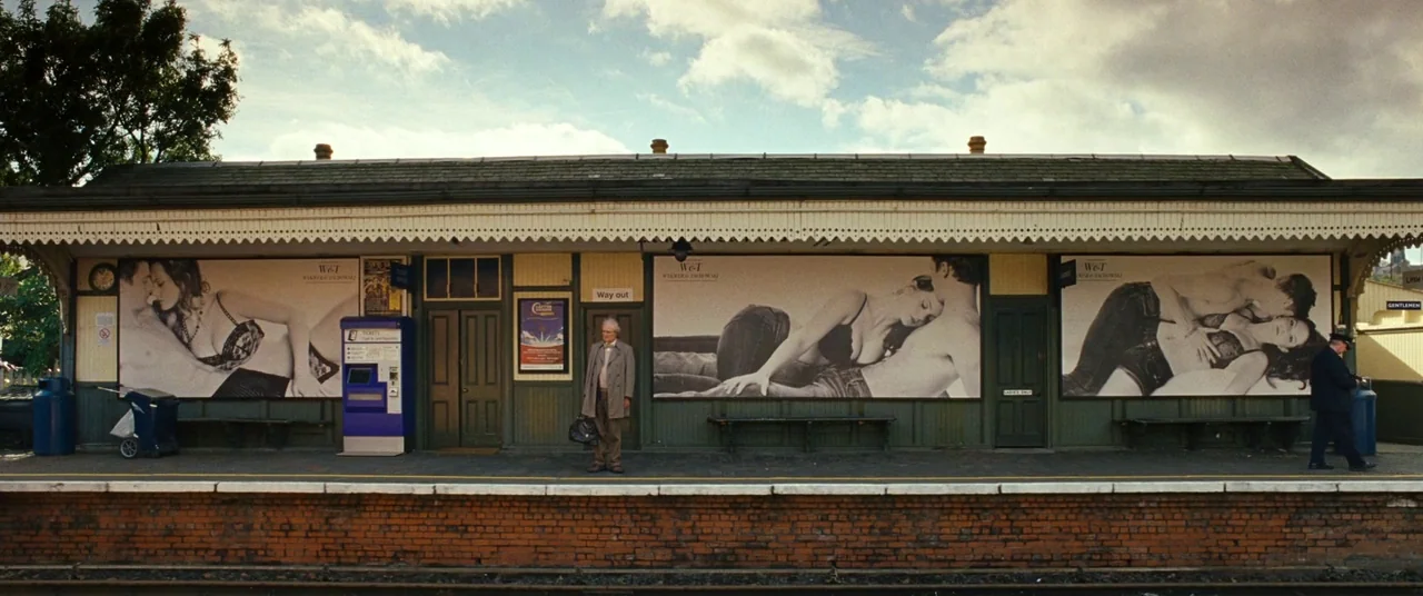 Cloud Atlas (2012) movie still: wide — A rural train platform featuring large black and white advertising posters on the walls. An elderly man in a…