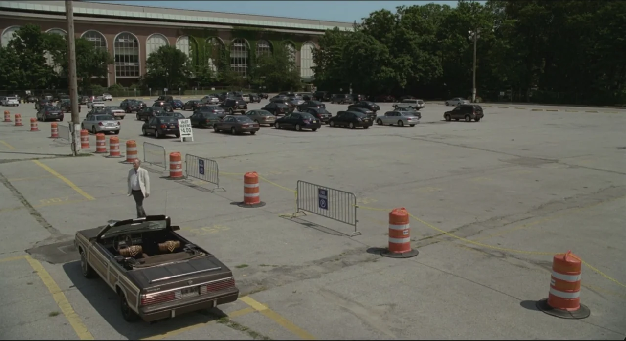 St. Vincent (2014) movie still: wide — A vast, paved parking lot features orange traffic barrels and metal crowd control fencing. An older man in a…