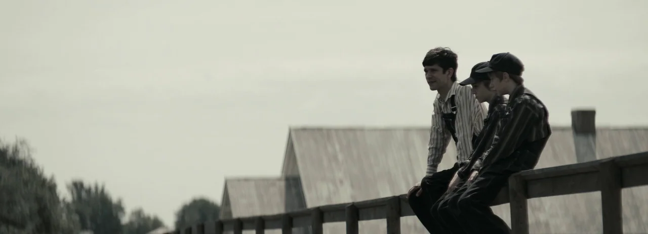 Women Talking (2022) movie still: wide — Three males sit side by side on a weathered wooden fence during daylight. In the distance stand several…