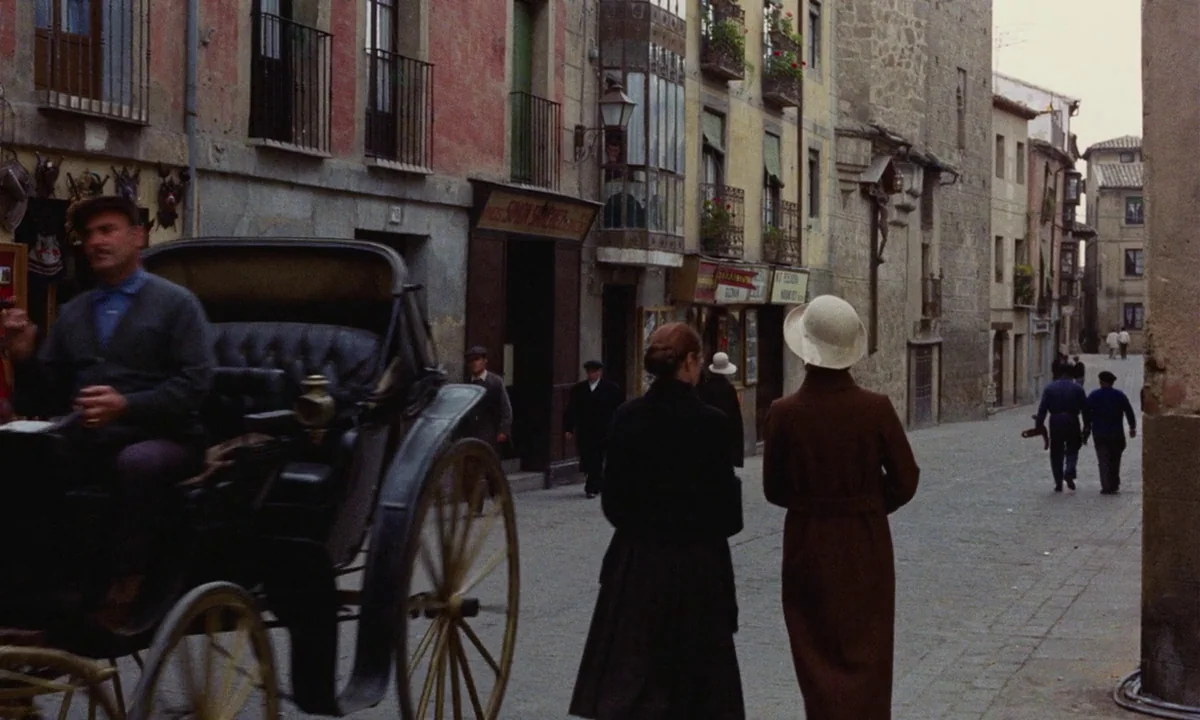 Tristana (1970) movie still: fullbody — A narrow European cobblestone street lined with multi-story stucco buildings and iron balcony railings. A…