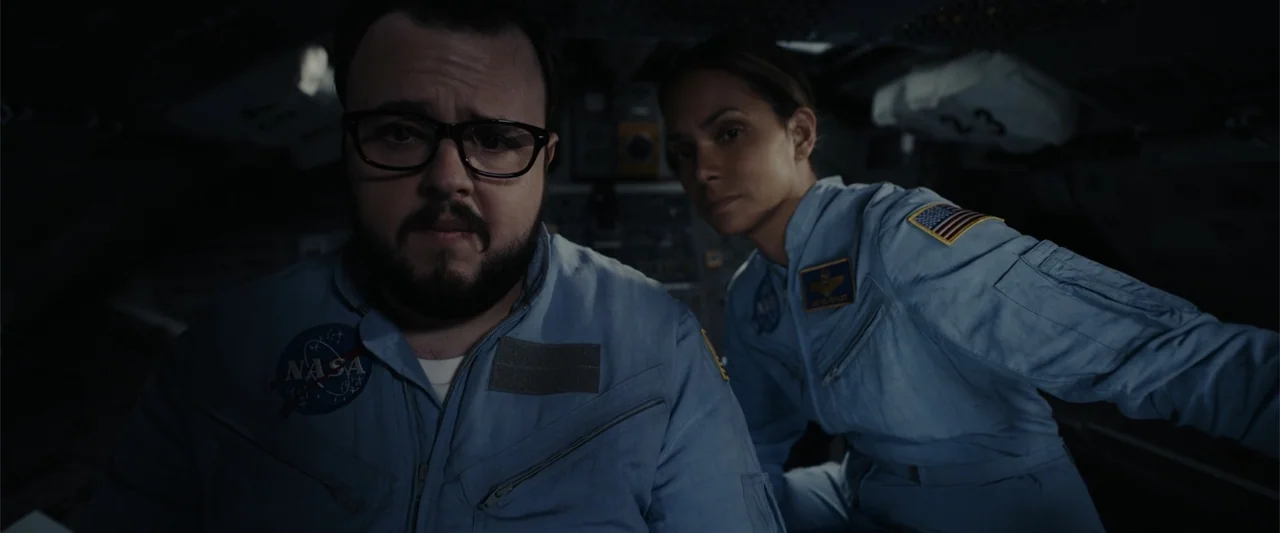Moonfall (2022) movie still: medium — A dark, cramped spacecraft cockpit contains instrument panels and buttons in the background. Two astronauts…