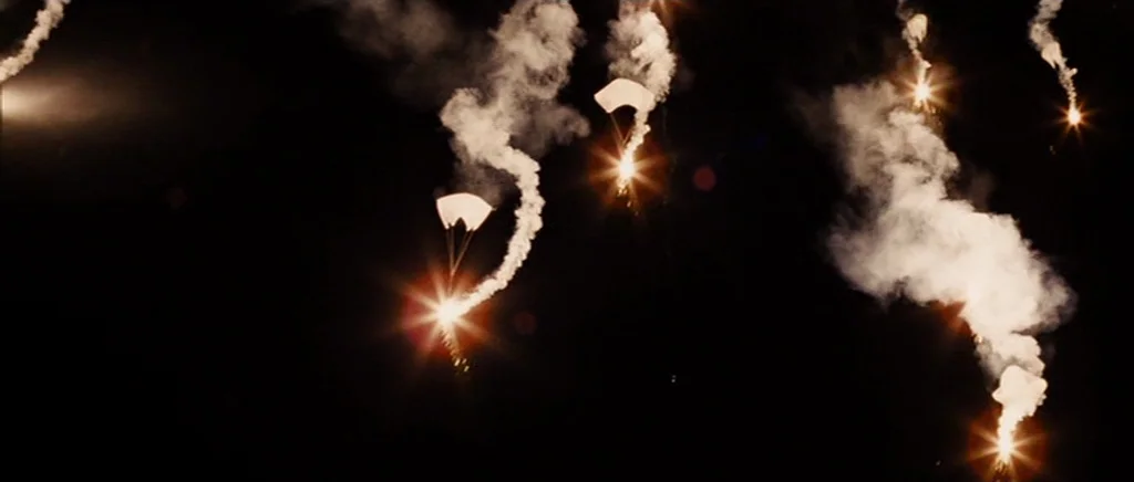 Jarhead movie still: wide — Several bright parachute flares drift slowly downward against an entirely dark night sky. Thick trails of white smoke…