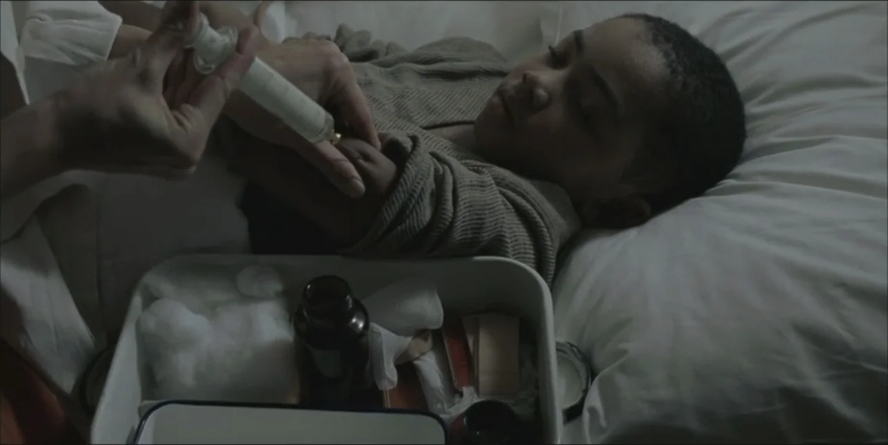 Exorcist: The Beginning (2004) movie still: medium — A person lying in a bed receives a medical injection from an unseen caregiver. A medical tray…