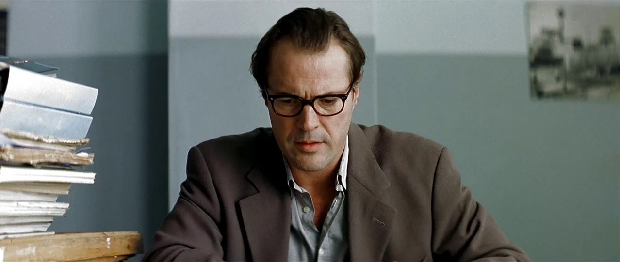 The Lives of Others (2006) movie still: medium — A man sits at a desk in a plain room with grey walls. He looks downward at paperwork while stacks…