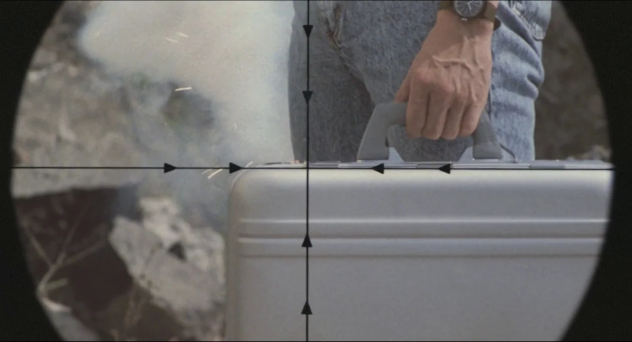 Godzilla vs. Biollante (1989) movie still: closeup — A close-up view through a sniper rifle scope reveals a metallic briefcase and jeans. Smoke…