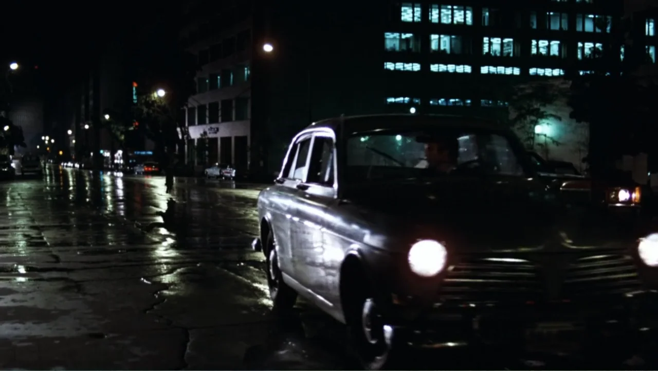 All the President's Men (1976) movie still: medium — A wet asphalt road at night reflects light from a parked vintage vehicle. A lone driver…