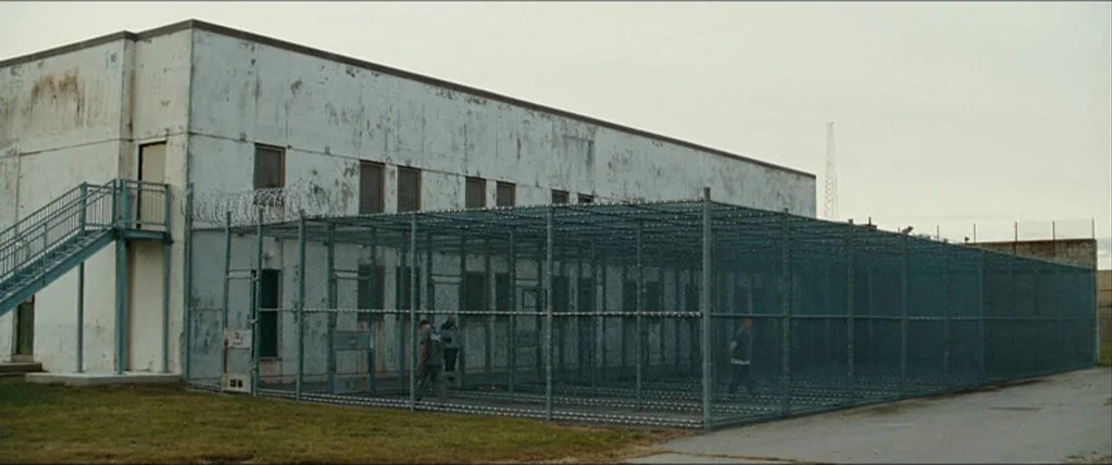 The Town (2010) movie still: wide — An institutional concrete building is adjoined by a large chain-link fenced recreation cage. Three individuals…