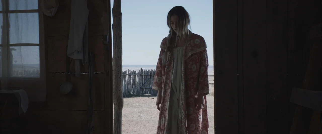 The Wind (2005) movie still: fullbody — A rustic wooden shack doorway opens onto a dry desert landscape with a fence. A young woman stands in the…