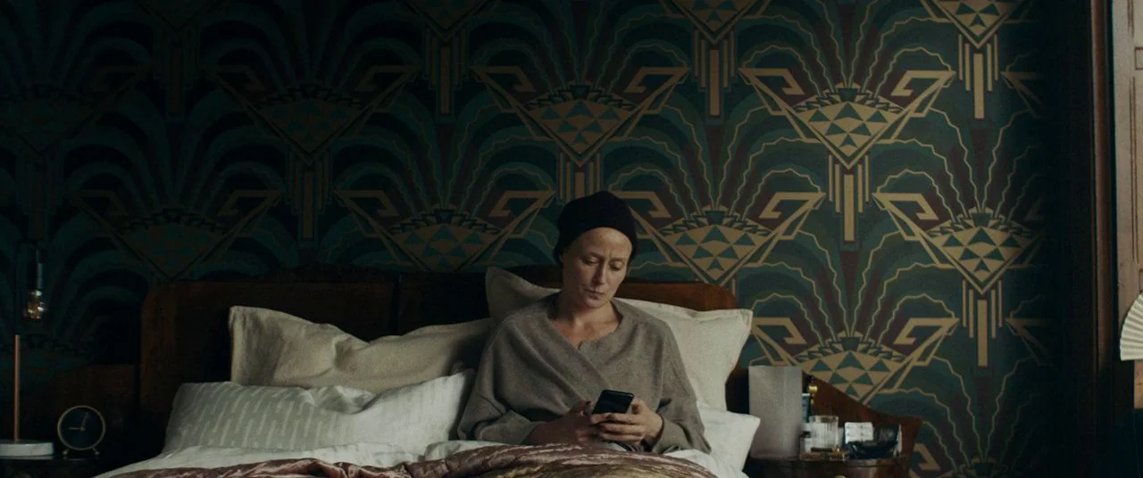 Saint Maud (2019) movie still: medium — A dimly lit room features distinctive geometric art deco patterned wallpaper. A person sits upright in a bed…