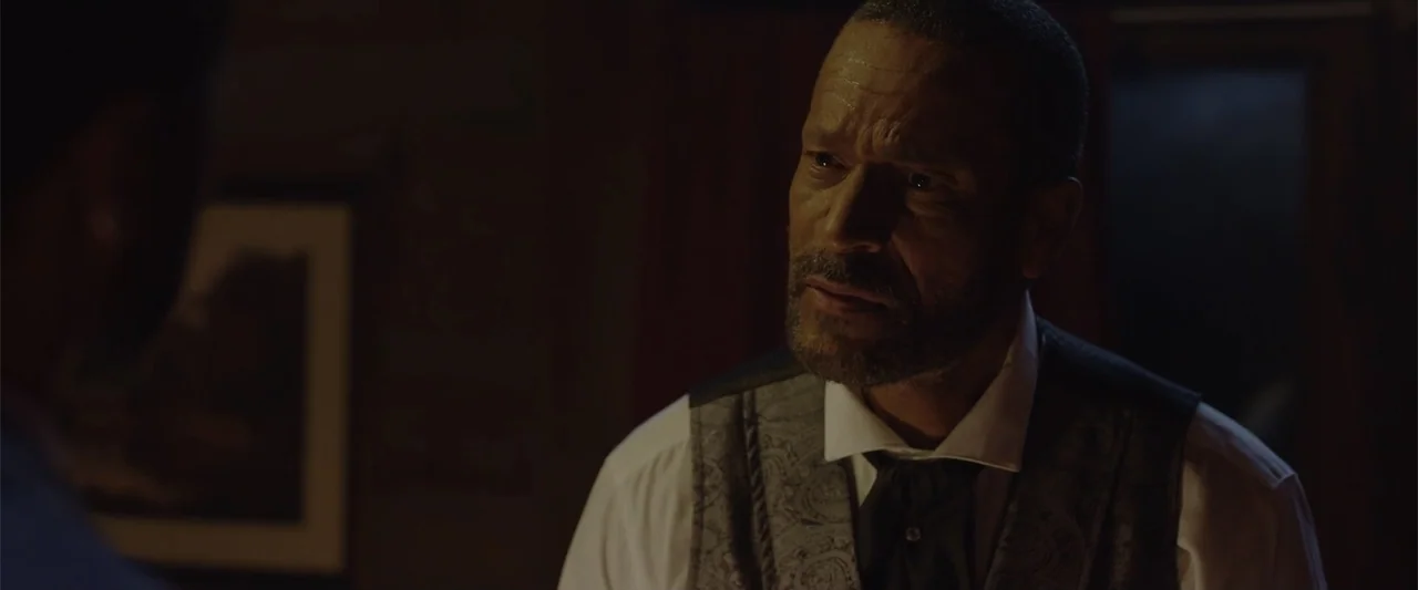 Ghosts of the Ozarks (2022) movie still: closeup — A dark indoor setting features a man in period clothing looking at another person. The man's face…