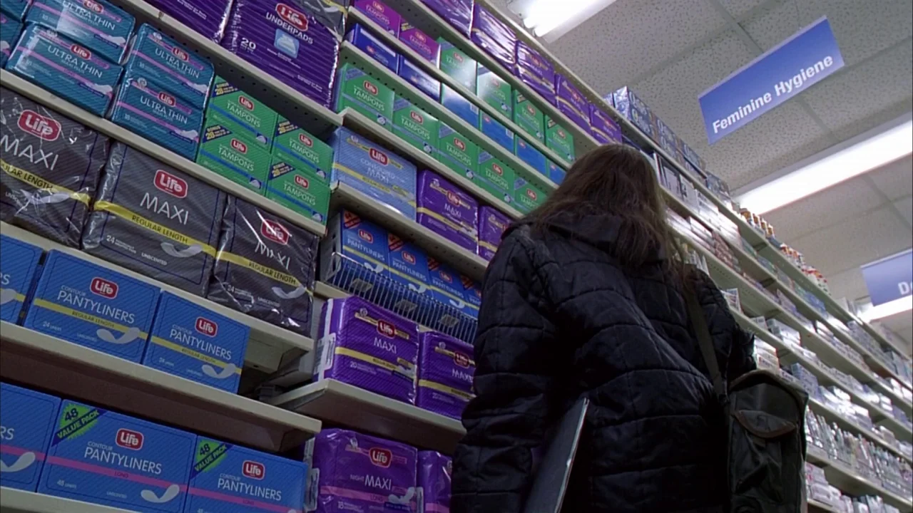 Ginger Snaps (2000) movie still: medium — A retail aisle stocked with colorful boxes of feminine hygiene products on metal shelving. A young woman…