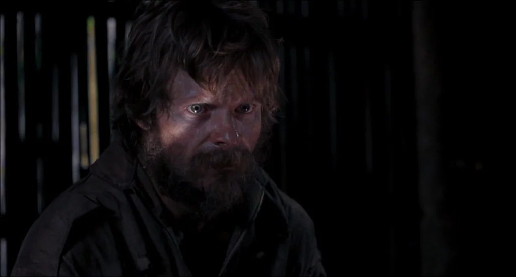 Rescue Dawn movie still: closeup — A man stands in a dimly lit, shadowed interior space with vertical wooden slats behind. He looks toward the side…