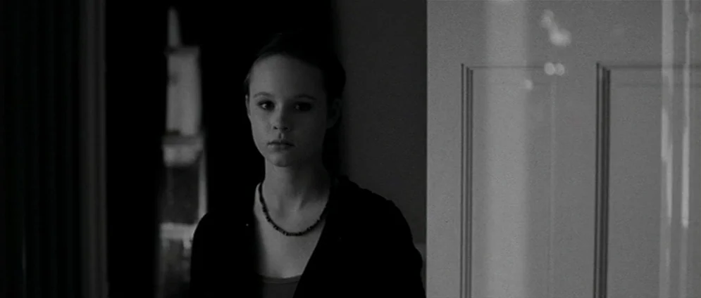 American Beauty (1999) movie still: medium — A dark interior doorway connects two rooms separated by a door frame. A young female stands inside the…
