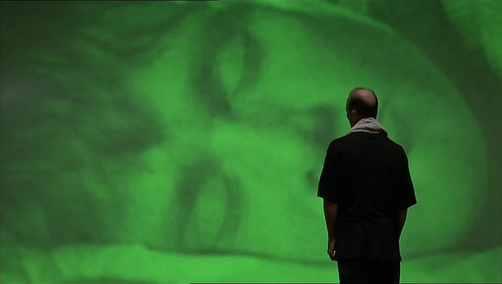 The Truman Show movie still: medium — A dark, empty space featuring a large projection of a human face in green. The projection is cast onto a flat…