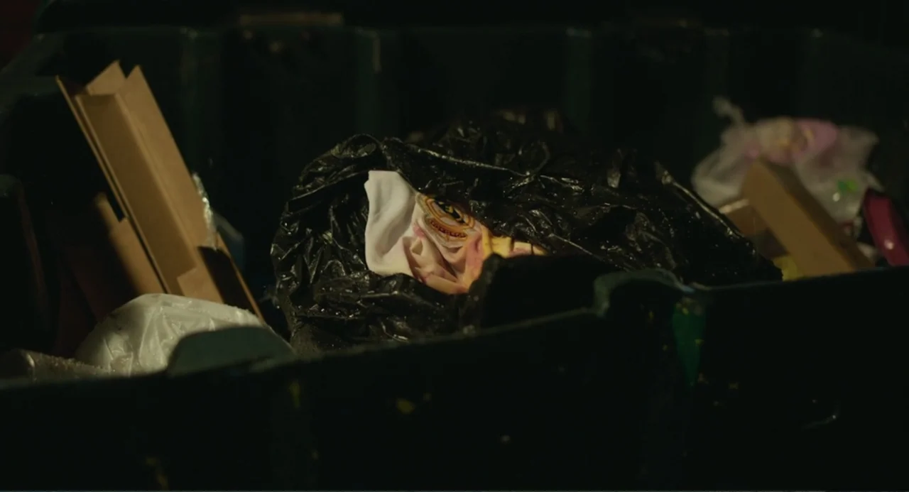 The Lift (2021) movie still: closeup — A dark plastic trash bin filled with discarded items and cardboard fragments. A piece of fabric with a sports…