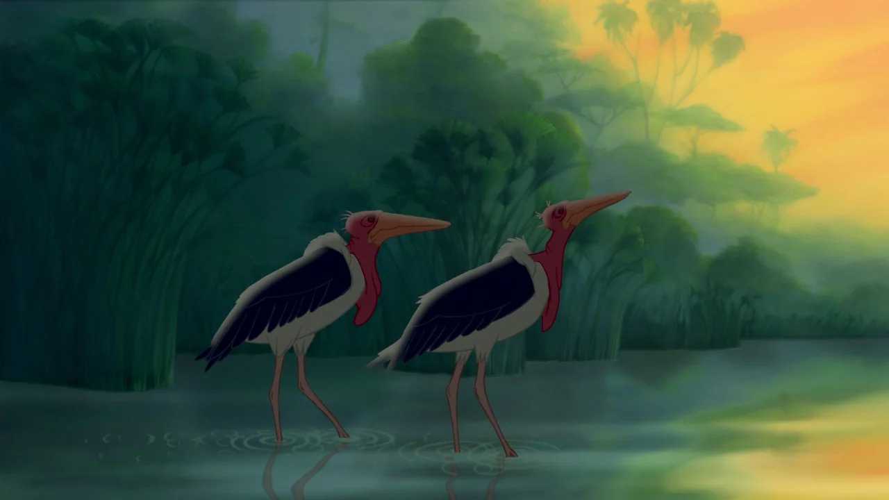 The Lion King (1994) movie still: medium — Two large birds stand in a shallow pool of water near dense green reeds. A vibrant orange sky suggests…
