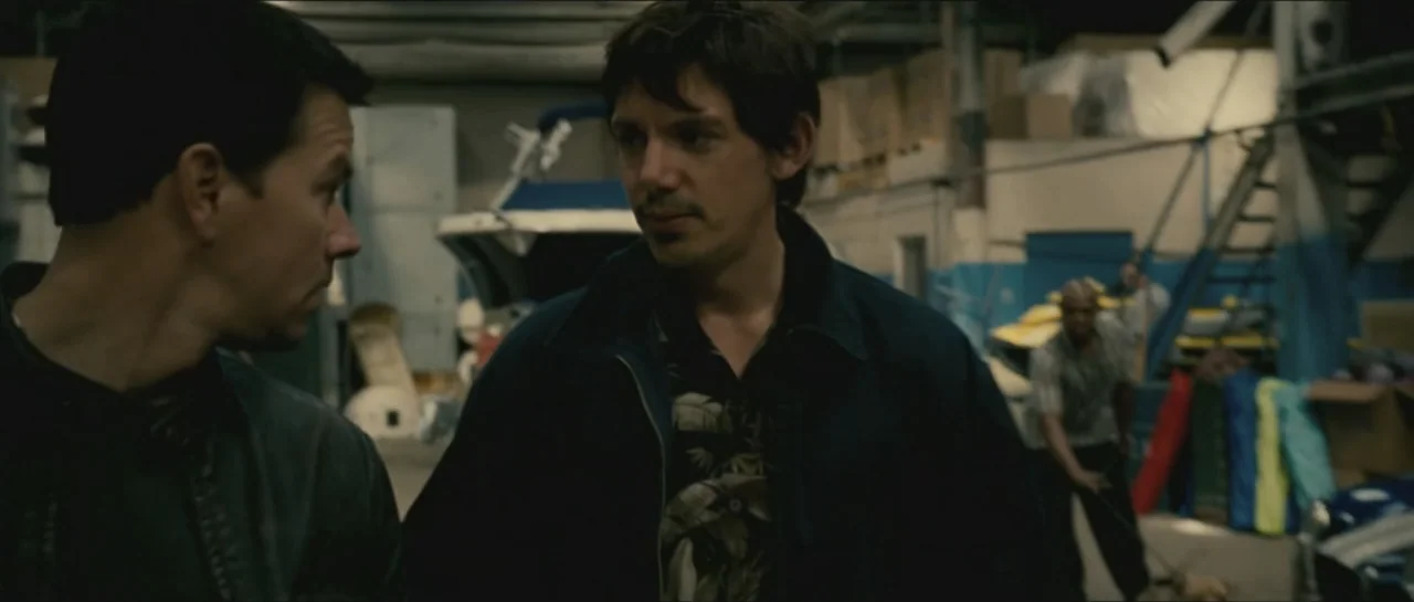 Contraband (2012) movie still: medium — An cluttered industrial auto repair shop contains various vehicles and metal scaffolding in back. Two men…