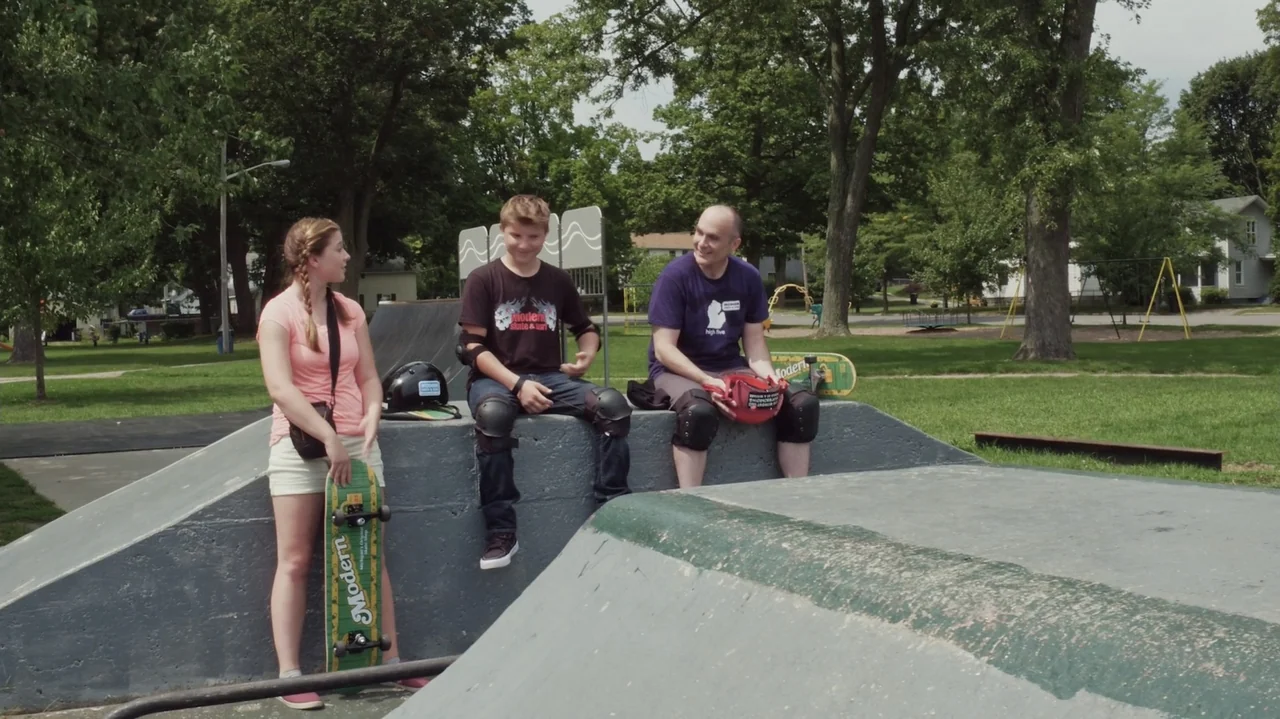 Sk8 Dawg (2018) movie still: fullbody — A public skatepark with concrete ramps and mature green trees in the background. Three people sit on a…