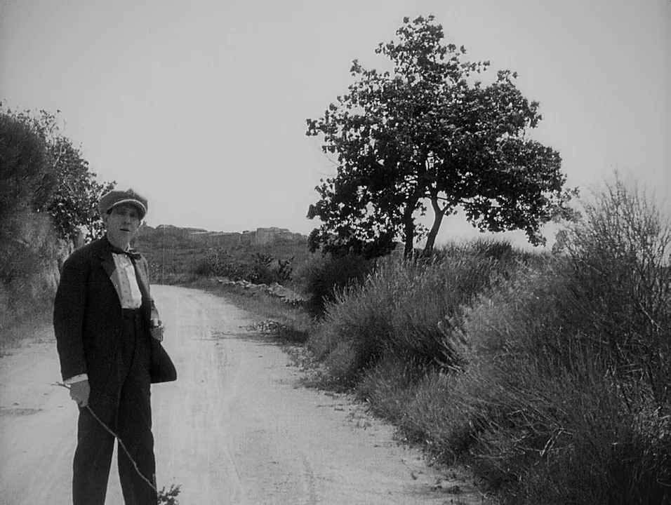 Coeur Fidele (1923) movie still: fullbody — A man stands on a rural dirt road lined with brush and a large tree. He holds a thin stick while looking…
