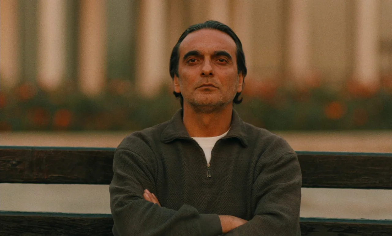 Taste of Cherry (1997) movie still: medium — A man sits on a wooden bench outdoors in front of a building facade. The subject stares directly at the…