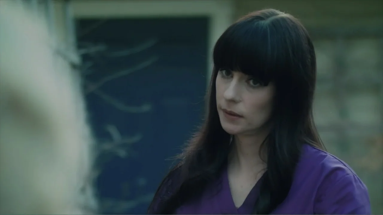 Storm Damage (2000) movie still: medium — An outdoor area with a blurred background showing a building exterior. A woman with dark hair in a purple…