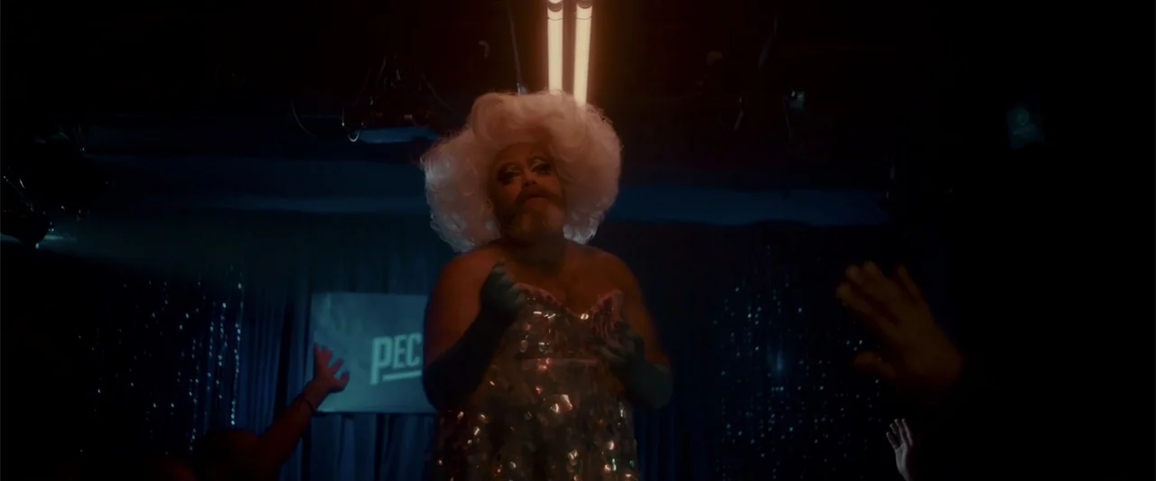 Jump, Darling (2022) movie still: medium — A dimly lit nightclub stage features a performer wearing a large blonde wig. The individual stands…