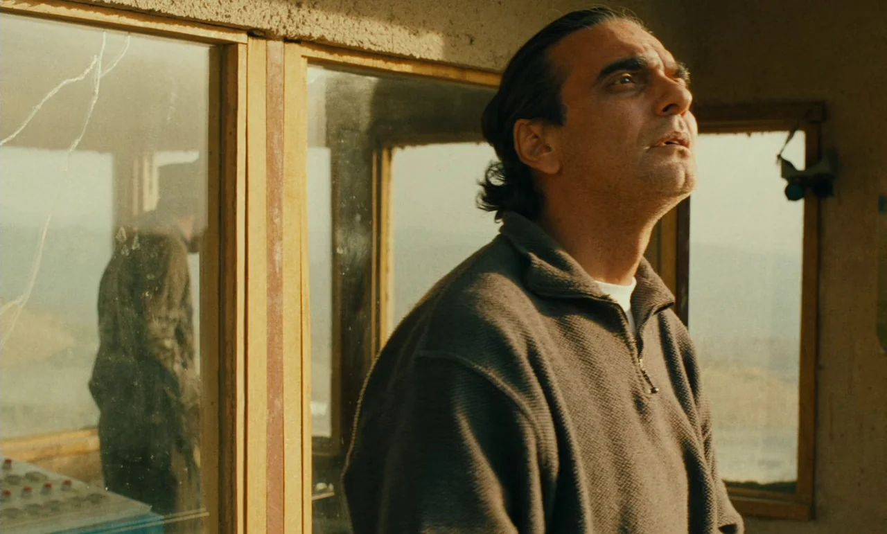 Taste of Cherry (1997) movie still: medium — A man stands inside a room with large glass panels overlooking a landscape. The middle-aged man looks…